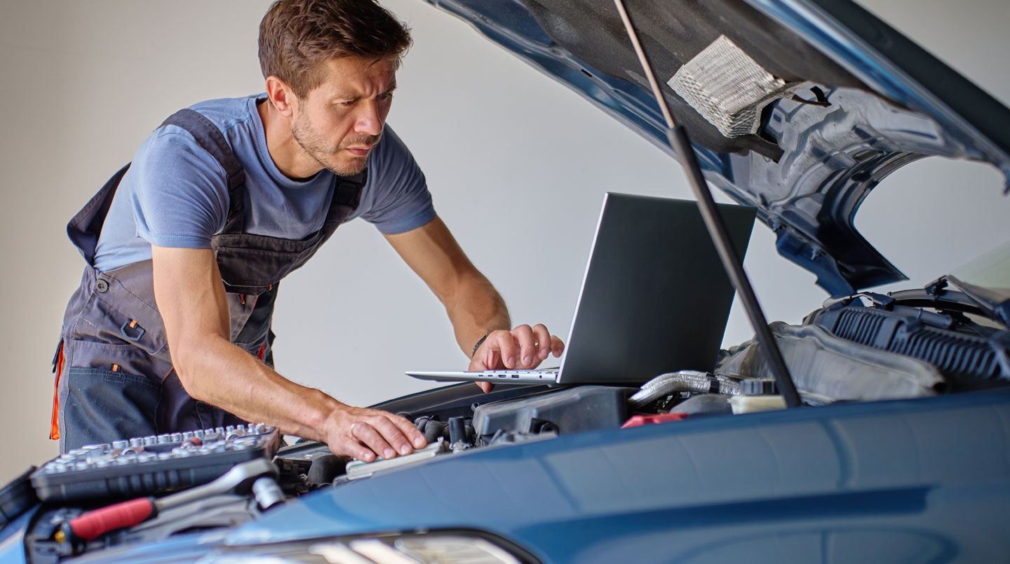 Mécanicien professionnel en bleu de travail inspectant le compartiment moteur ouvert d'une berline d'occasion dans un atelier automobile, carnet d'entretien et tablette de diagnostic posés sur l'aile du véhicule